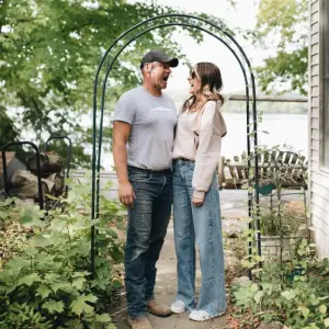 A man and woman share a laugh under a garden archway. The man wears a cap, t-shirt, and jeans, while the woman is in a light blouse and wide-leg jeans. They stand amidst green foliage with a lake and wooden chairs in the background.